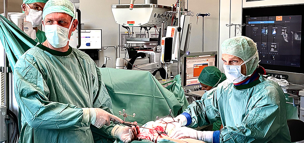 Surgeons in green scrubs and masks perform surgery in an operating room with monitors and equipment.